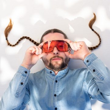 Scoobert Doobert reclining in red wraparound sunglasses, two long braids fanned out behind his head.